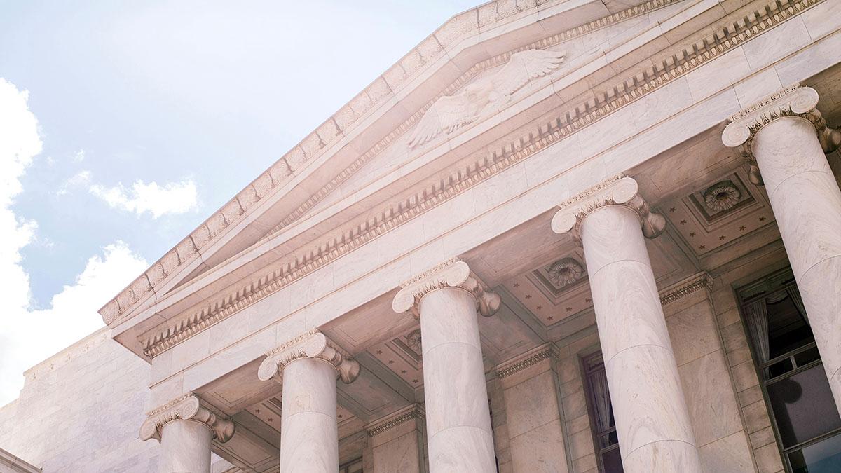 A close-up photo of the front of a grand building. The building has a row of tall, white columns with ornate details at the top. There is a clear blue sky in the background.