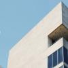 Modern architectural building with clean lines and a minimalist design. The structure features light-colored stone walls and large glass windows, set against a clear blue sky. The design includes sharp angles and an extended balcony area.
