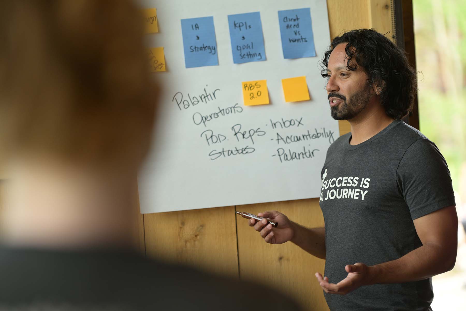 Person standing at a board with post it notes on it. He is wearing a t-shirt that says, "Success is a Journey" is
